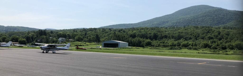 The view from the airport ramp. The airplane is parked in the foreground, with a hanger behind and a tall mountain in the background.
