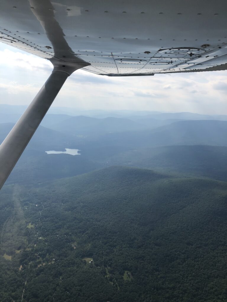 A view out the right of the plane in flight. The Catskill mountains are in view, with a small lake also visible.